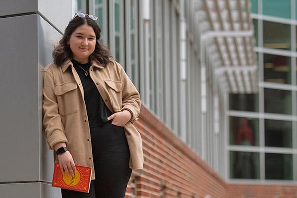 English student leaning against the Student Engagement Center building while holding a novel